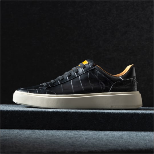 A side view of a men’s dress sneaker In black croco leather with the toe facing the left. Vue de côté d'une basket habillée pour homme en cuir croco noir avec le bout tourné vers la gauche.