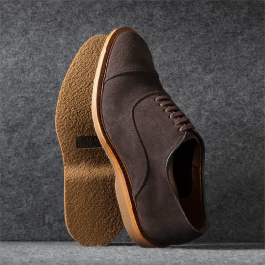 A pair of brown suede cap toe oxford dress shoes displayed vertically, with one sole facing forward. Une paire de chaussures habillées Oxford à bout droit en suède brun, présentées verticalement, avec une semelle tournée vers l'avant.