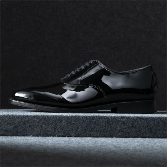 A side view of a black patent leather oxford dress shoe with the toe facing left. Vue latérale d'une chaussure Oxford en cuir verni noir avec l'orteil tourné vers la gauche.